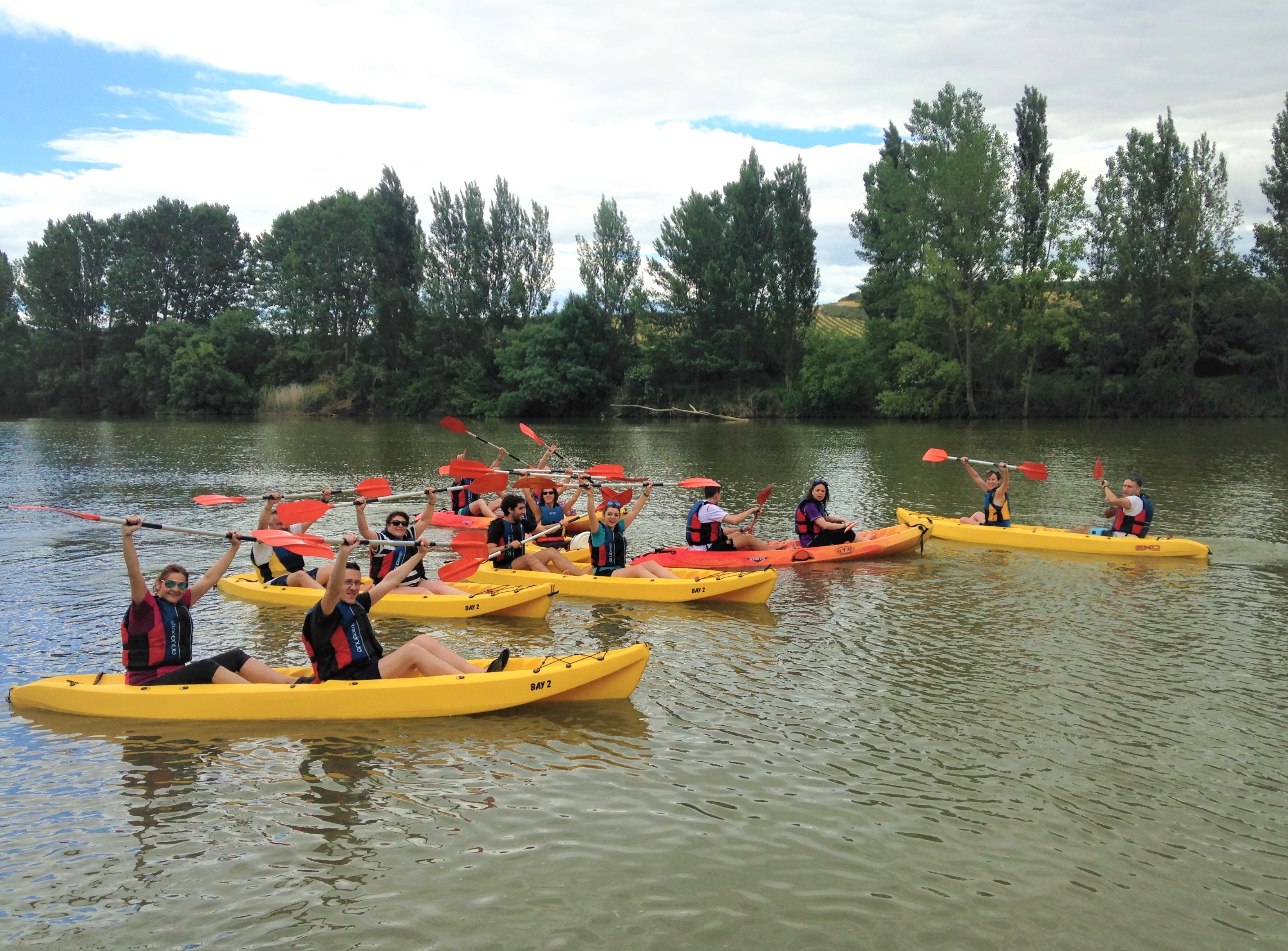 un grupo de unas 20 personas disfrutando de un paseo en kayak por el rio ebro montados en caoas amarillas y levantando los brazos en forma de alegría
